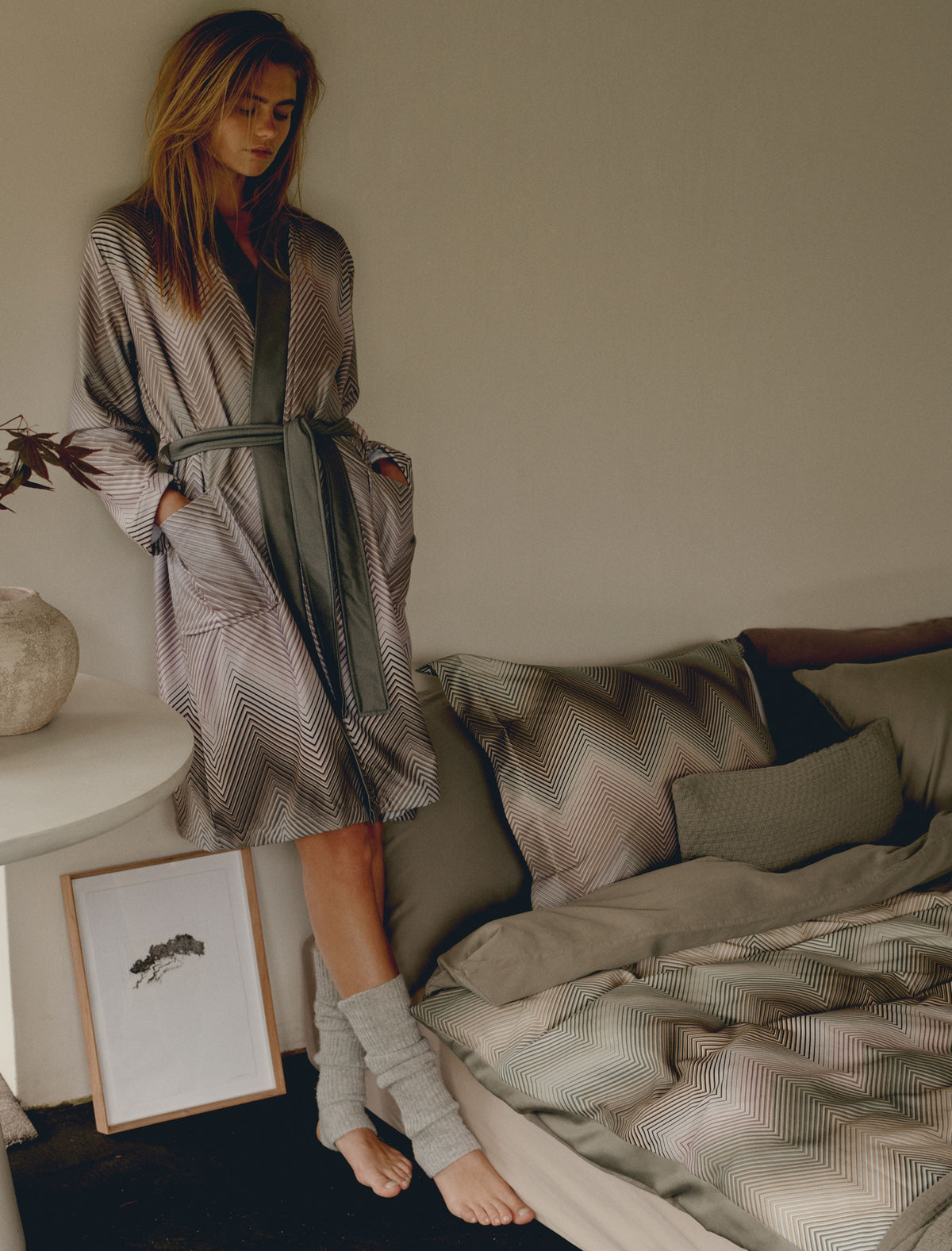 Een vrouw met een badjas aan in zigzag print die naast een bed staat met een dekbedovertrek in zigzag print in groene kleuren