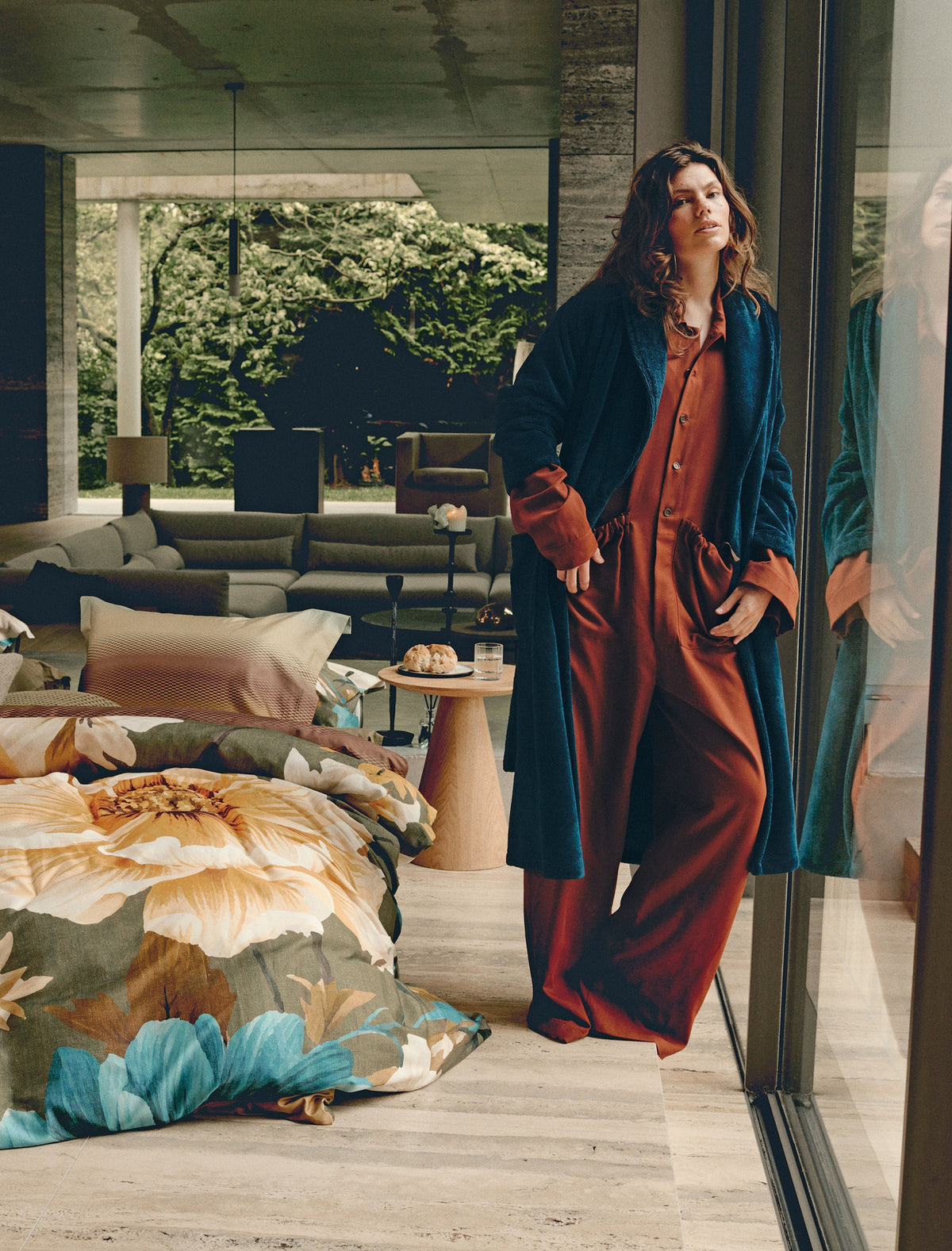 Een vrouw met een groene badjas aan die naast een bed staat met een dekbedovertrek met bloemen print in gele, blauwe en groene kleuren