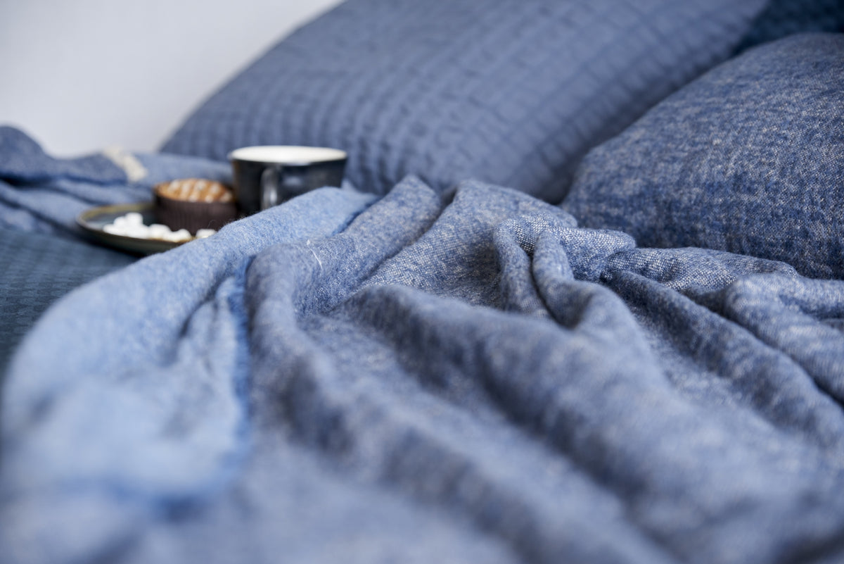 detail shot van een plaid in een jeans blue kleur wat op een bed ligt met een kopje koffie en ontbijt