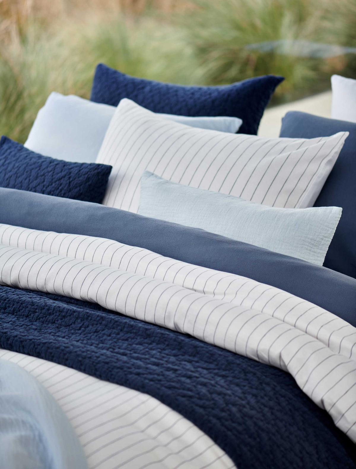 detail shot van een bed met een wit dekbedovertrek met dunne blauwe strepen en en dekbedovertrek met wafelstructuur in een jeans blauwe kleur. Op het bed ligt een sierkussen in een licht blauwe kleur