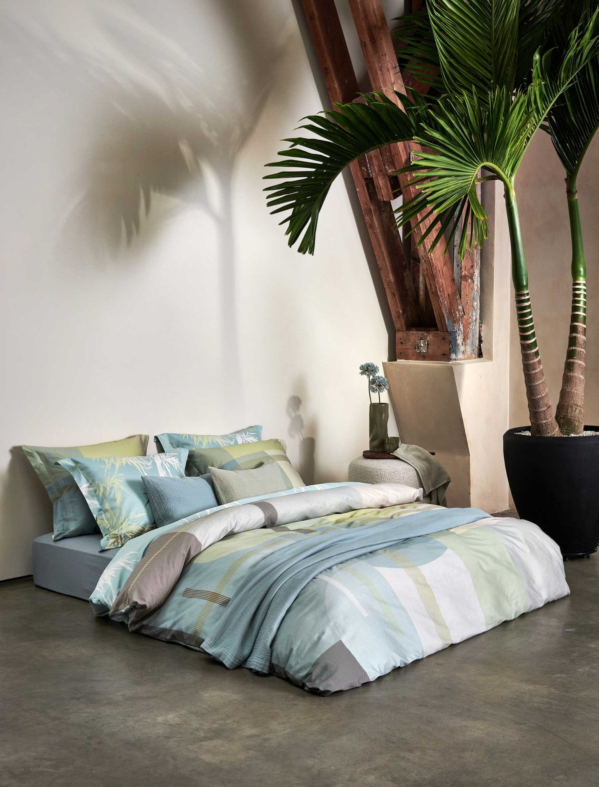 Palm Paradise thyme dekbedovertrek heeft silhouetten van palmbomen in bruin, beige, groen en wit op een lichtblauwe achtergrond. De achterkant van het dekbedovertrek heeft dezelfde print. De kussens hebben aan beide kanten palmbomen.