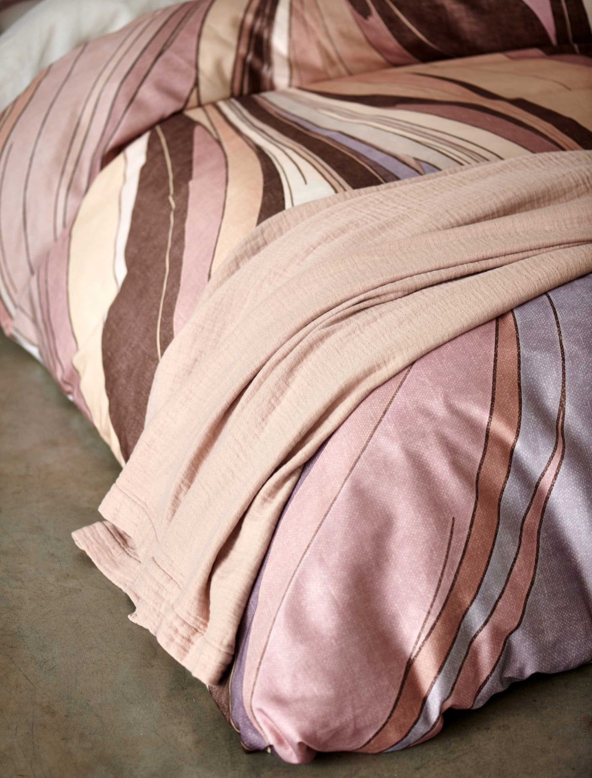 Swirls moonscape dekbedovertrek heeft een abstracte versie van een marmerpatroon met paars, roze en bruine tinten. De achterkant van het dekbedovertrek is effen offwhite. De kussens hebben aan beide zijden hetzelfde patroon. 