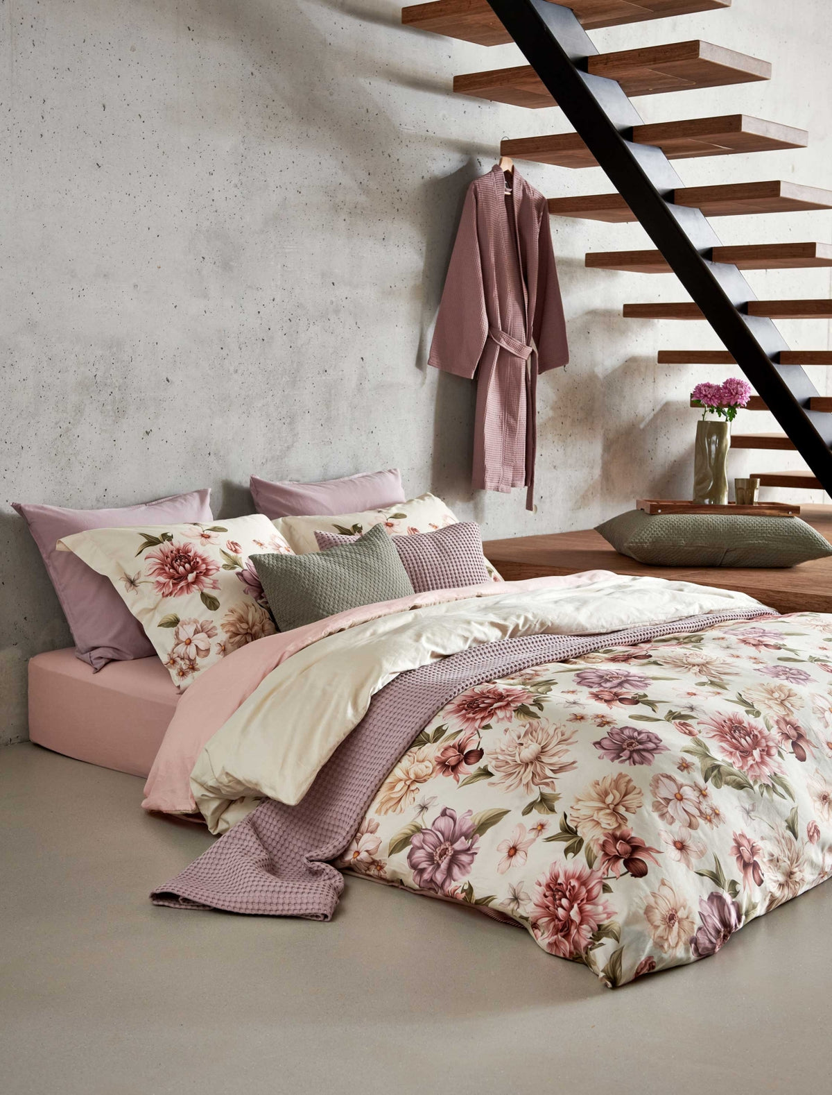 Een bed in een rustige setting met een dekbedovertrek met rode, paarse en beige bloemen. Op het bed ligt een sprei in een lila kleur en in een groene kleur