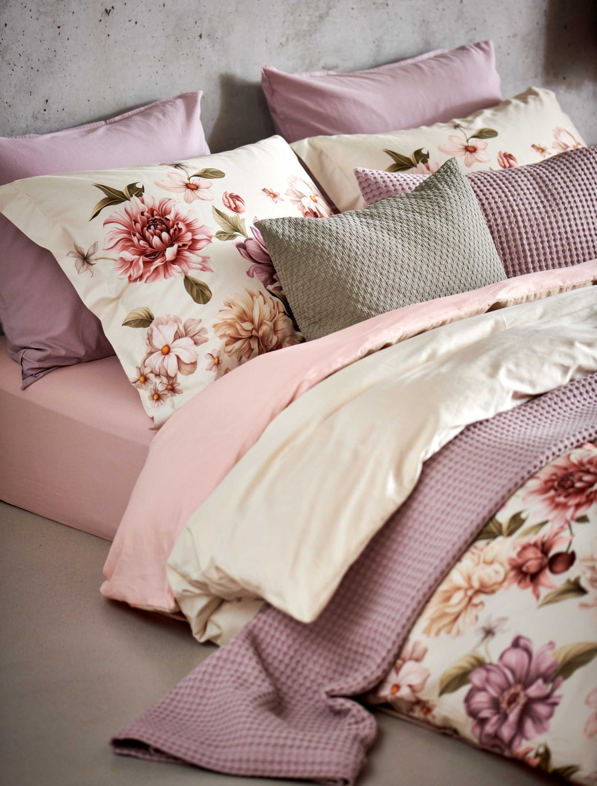 detail shot van een dekbedovertrek in een beige kleur met roze, paarse en beige bloemen op het bed ligt een sprei en een sierkussen in een paarse kleur en een sierkussen in een groene thyme kleur